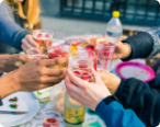 People holding glasses filled with something to drink toasting while eating outside.