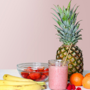 Fruit in front of a pink background on a white table with a pink smoothie in front of a pineapple.