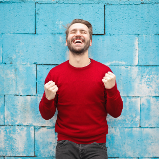 Blue brick background with a man smiling in a red shirt with their hands in fists.